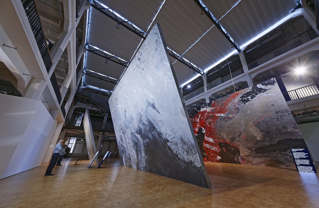 A person standing in front of a huge canvas on which a total of a cracked ground can be seen