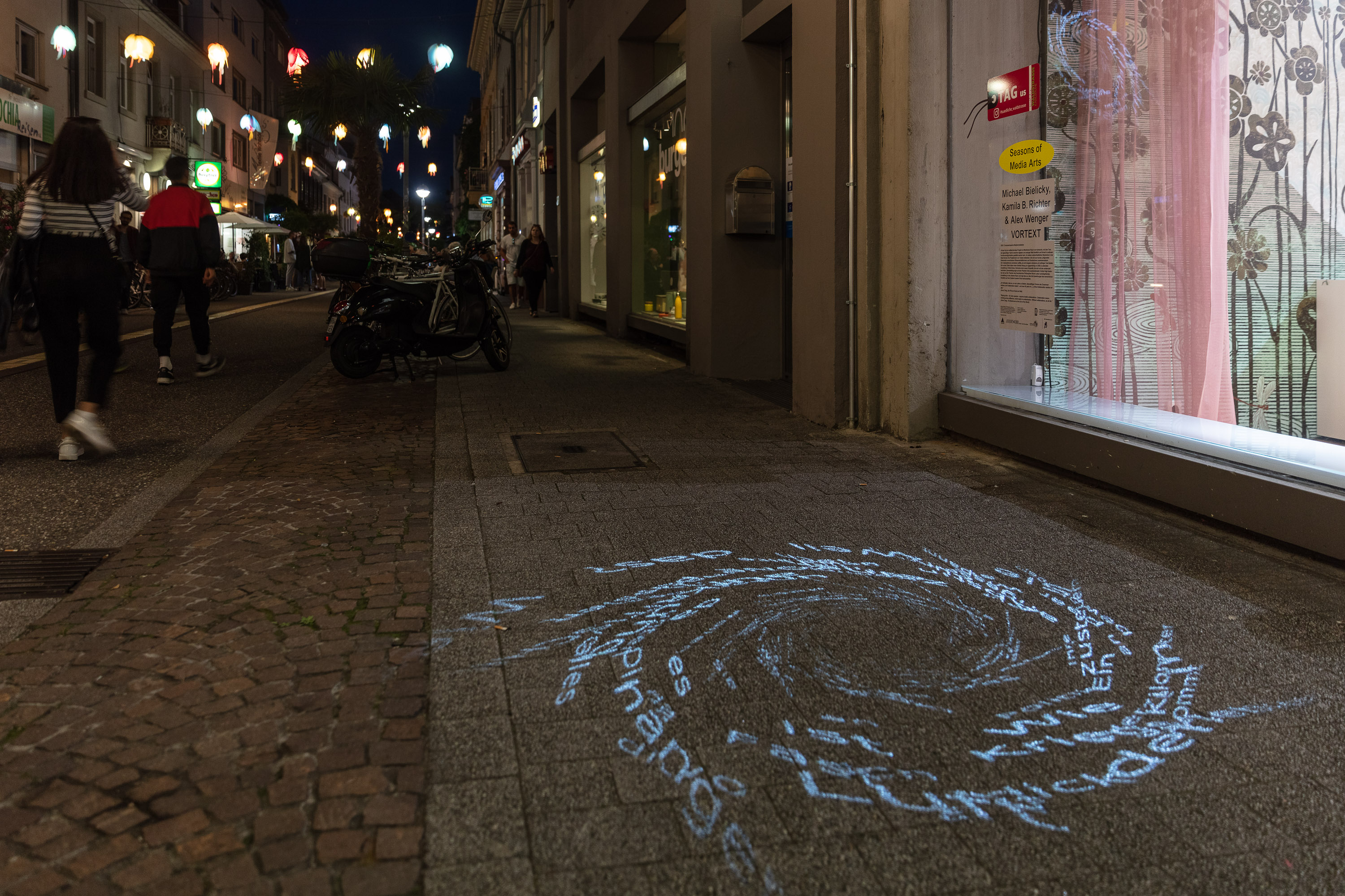 Zu sehen ist ein dunkler Raum, der die Form einer Röhre annimmt. Die Wände zeigen Lichtprojektionen, die aus einzelnen Buchstaben bestehen.