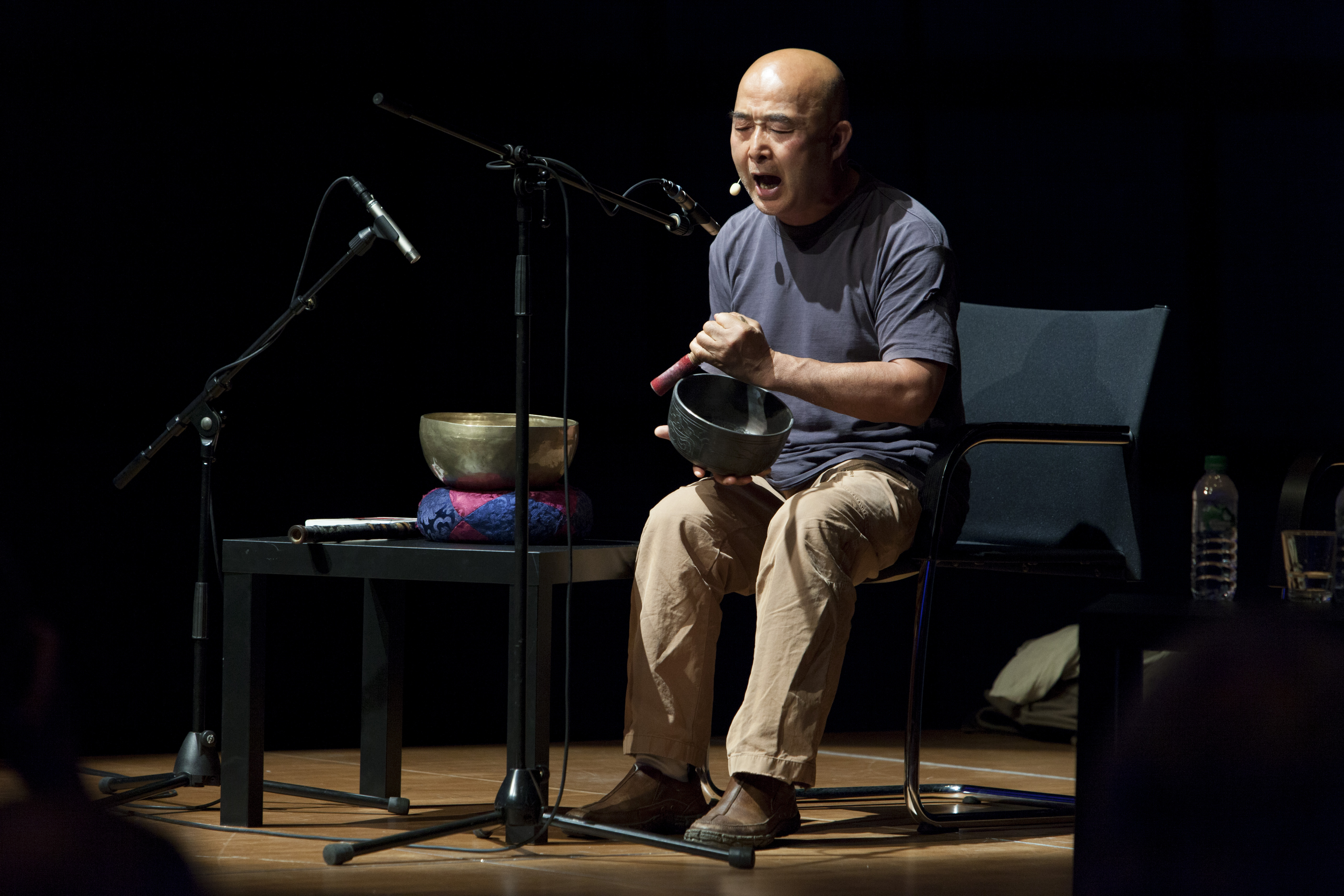 A man is sitting on a stage, making music with a  sound bowl while he is singing