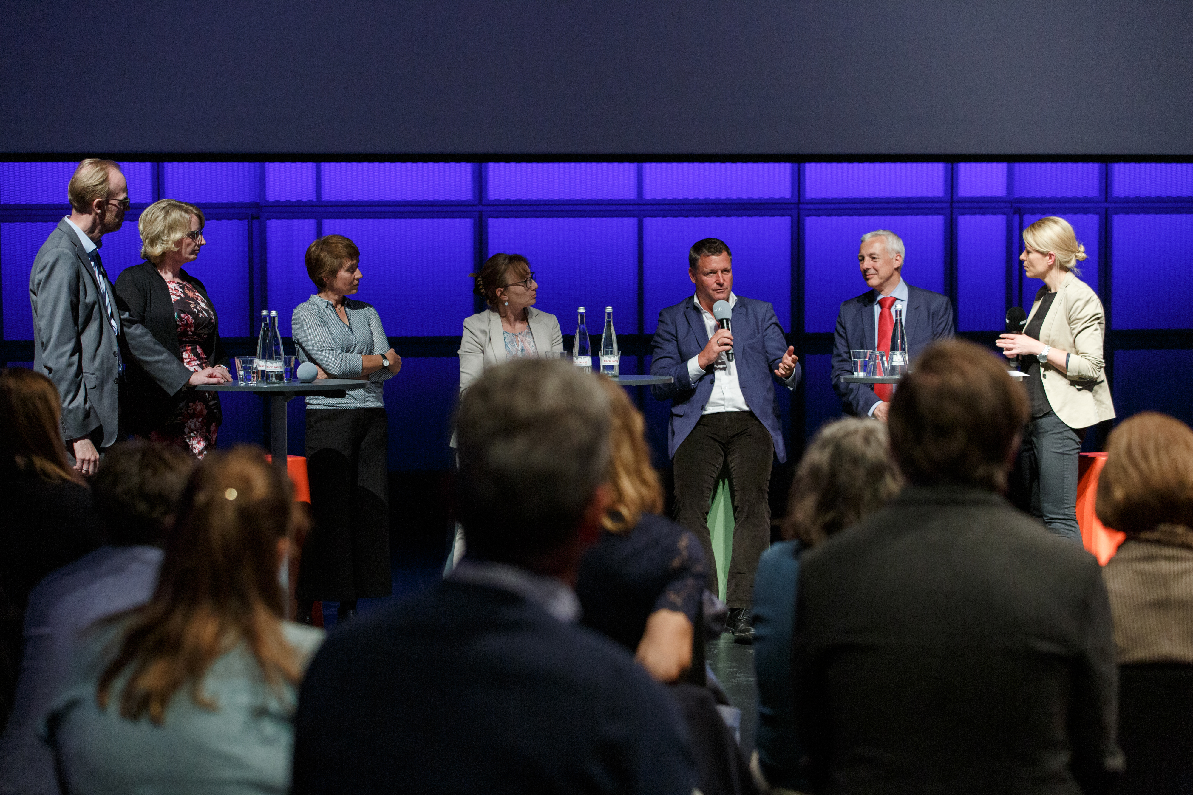 Vor einer Gruppe Menschen diskutieren sieben Personen an Stehtischen mit Mikrofonen in der Hand.