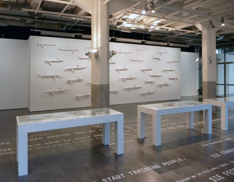 View of the exhibition »Symbiosis«: In the foreground, white cabinets, in the background on the wall: white guns