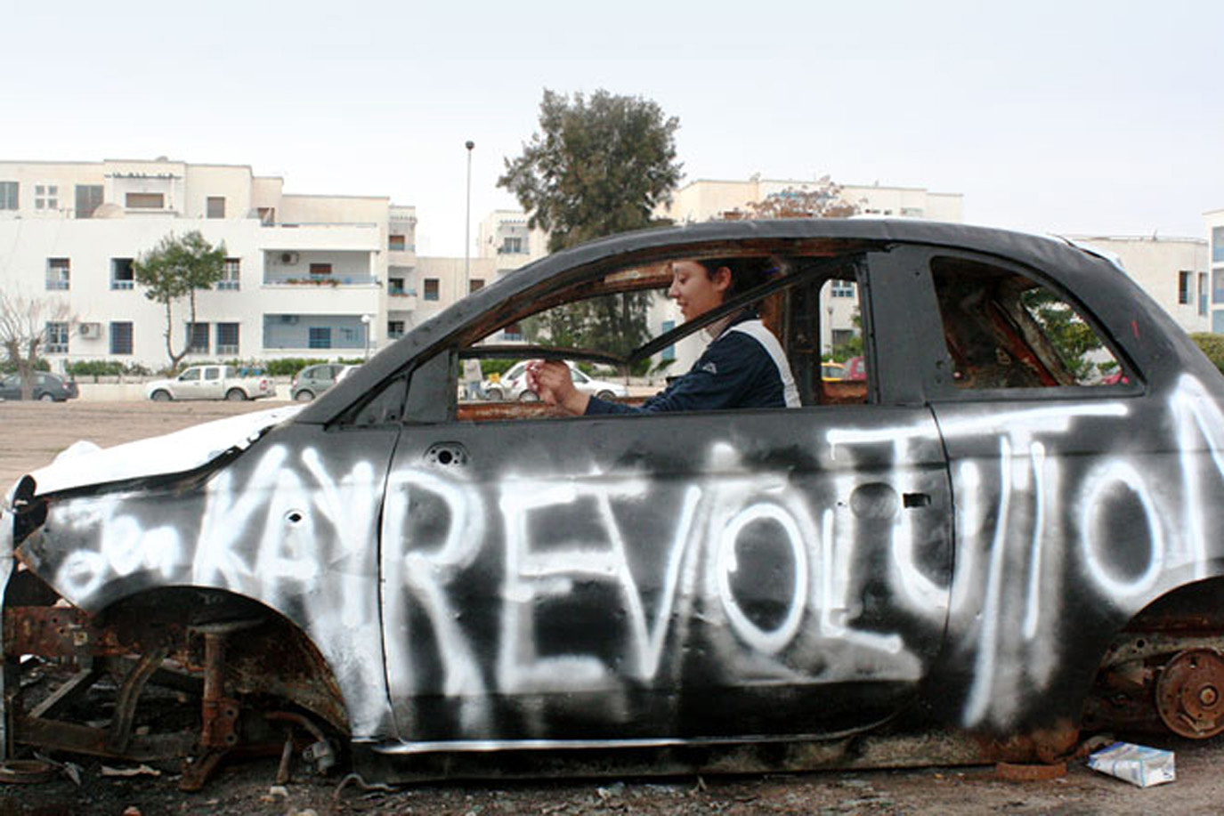 Eine Frau sitzt in einem funktionsuntüchtigen Auto. Auf der Fahrerseite wurde mit weißer Farbe »Revolution« gesprüht.