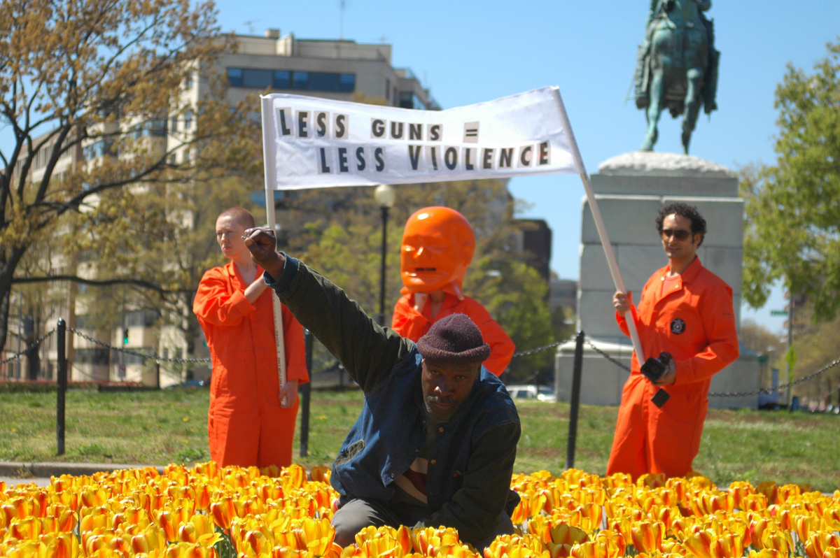 Ein Mann im Blumenbeet hebt seine rechte Faust als Zeichen des Protests. Hinter ihm drei Männer mit einem Transparent.