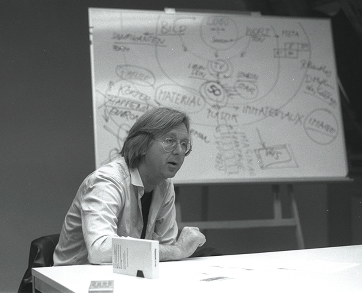 Black and white photograph of a man sitting at a table. In the background a flip chart.