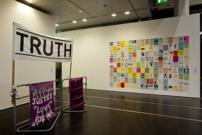 Auf der linken Bildhälfte sind drei Transparente zu sehen. Auf einem steht 'Truth'. Auf der rechten Bildhälfte kleben an einer Wand viele Papiere. Sie sind farbig und mit Texten versehen. Sie bilden in ihrer Masse eine großes Rechteck.