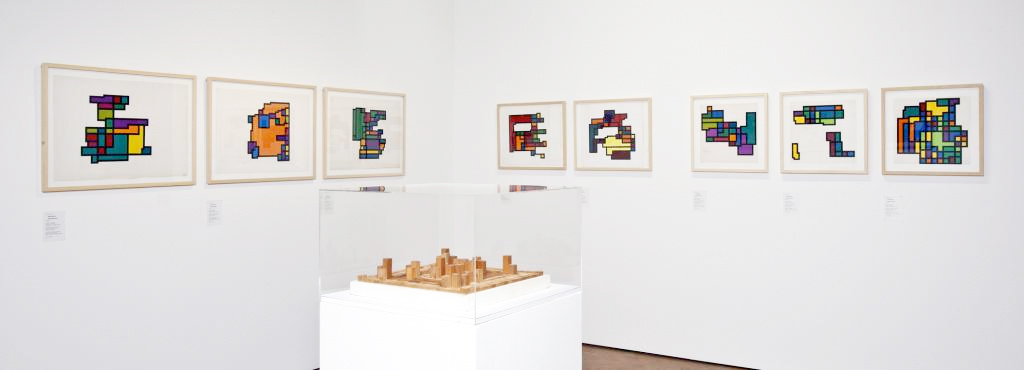Ein Blick in die Ausstellung »Hiroshi Kawano«: Ein Raum mit sieben Gemälden mit geometrischen Mustern, in der Mitte eine abstrakte Holzskulptur.