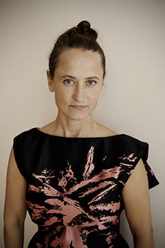 Halbportrait einer Frau. Sie trägt die Haare geschlossen. Ihr schwarzes ärmelloses Oberteil hat einen altrosa farbenen abstrakten Aufdruck.