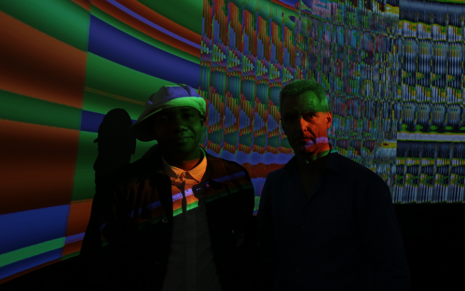 Two men standing in a dark room in front of a screen with colorful colors.
