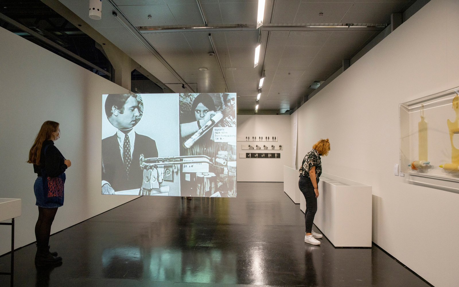 Zwei junge Frauen im Ausstellungsraum. Eine steht an der linken Wand und die andere schaut an der rechten Wand in Schaukästen. Zwischen ihnen spannt sich eine Leinwand, auf der ein Filmausschnitt mit Menschen zu sehen ist.