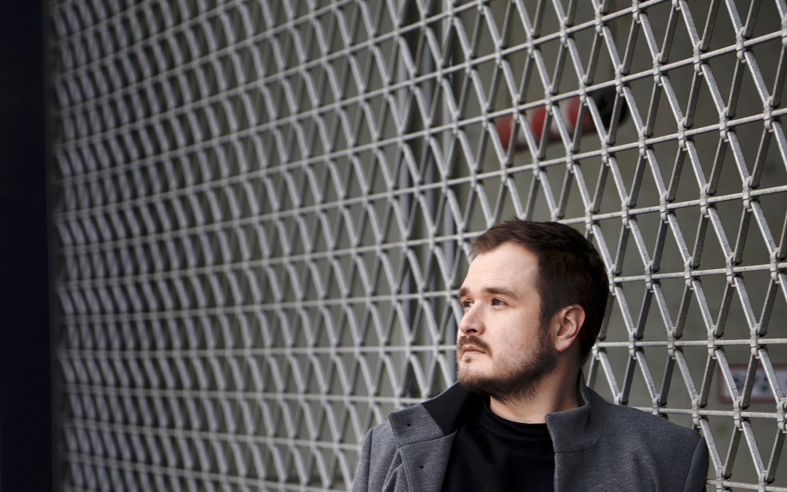 Foto von Artur Dziuk mit grauem Mantel und schwarzen Haaren. Er lehnt an einem Maschendrahtzaun.