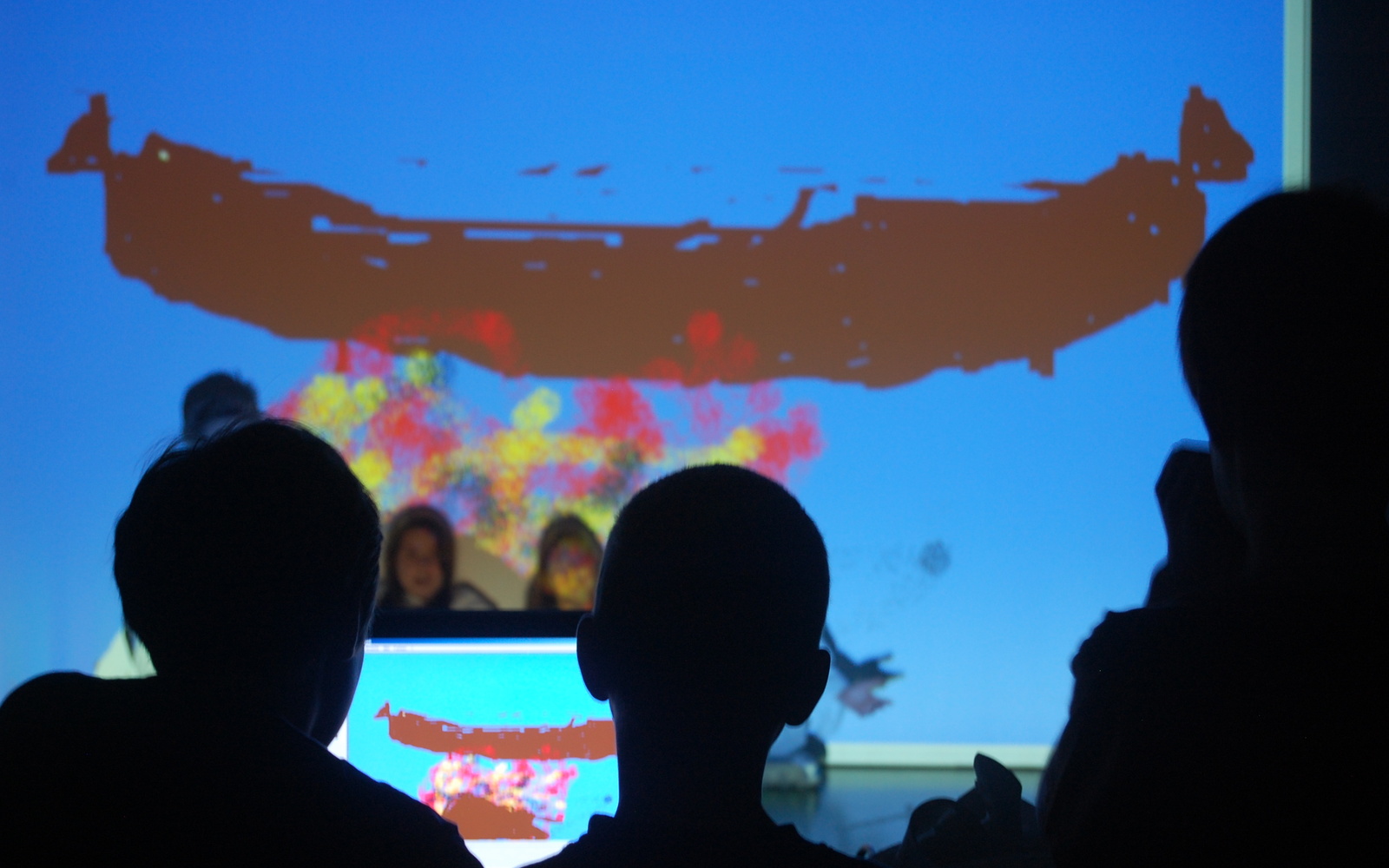Kinder sitzten vor dem Computer und bearbeiten ein Bild, das mit dem Beamer auf eine Leinwand und die Anzüge der Kinder davor geworfen wird. Sie sitzen unter einer großen Wurst.