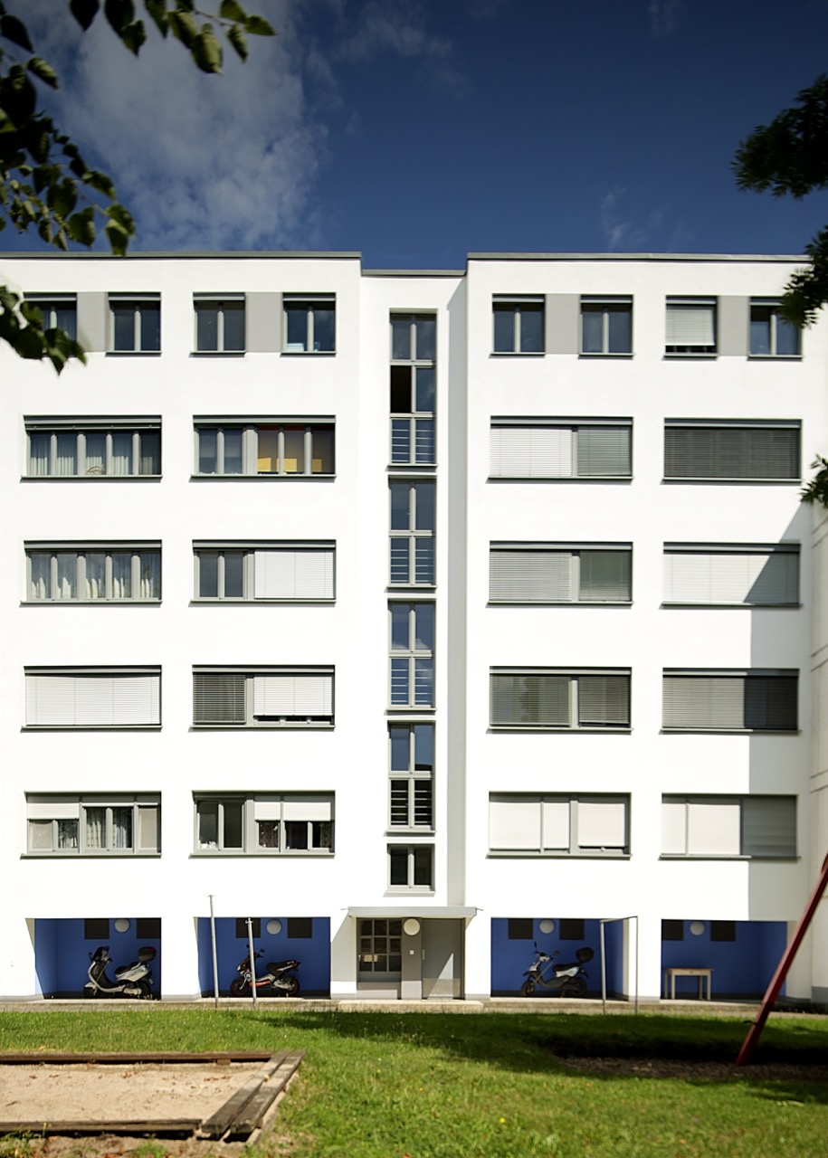 Zu sehen ist ein großes Weiße Gebäude mit einer Galerie im Erdgeschoss, deren Wände blau gestrichen sind. Das Gebäude befindet sich in der Dammerstocksiedlung in Karlsruhe, die aus der Bauhaus Zeit stammt.