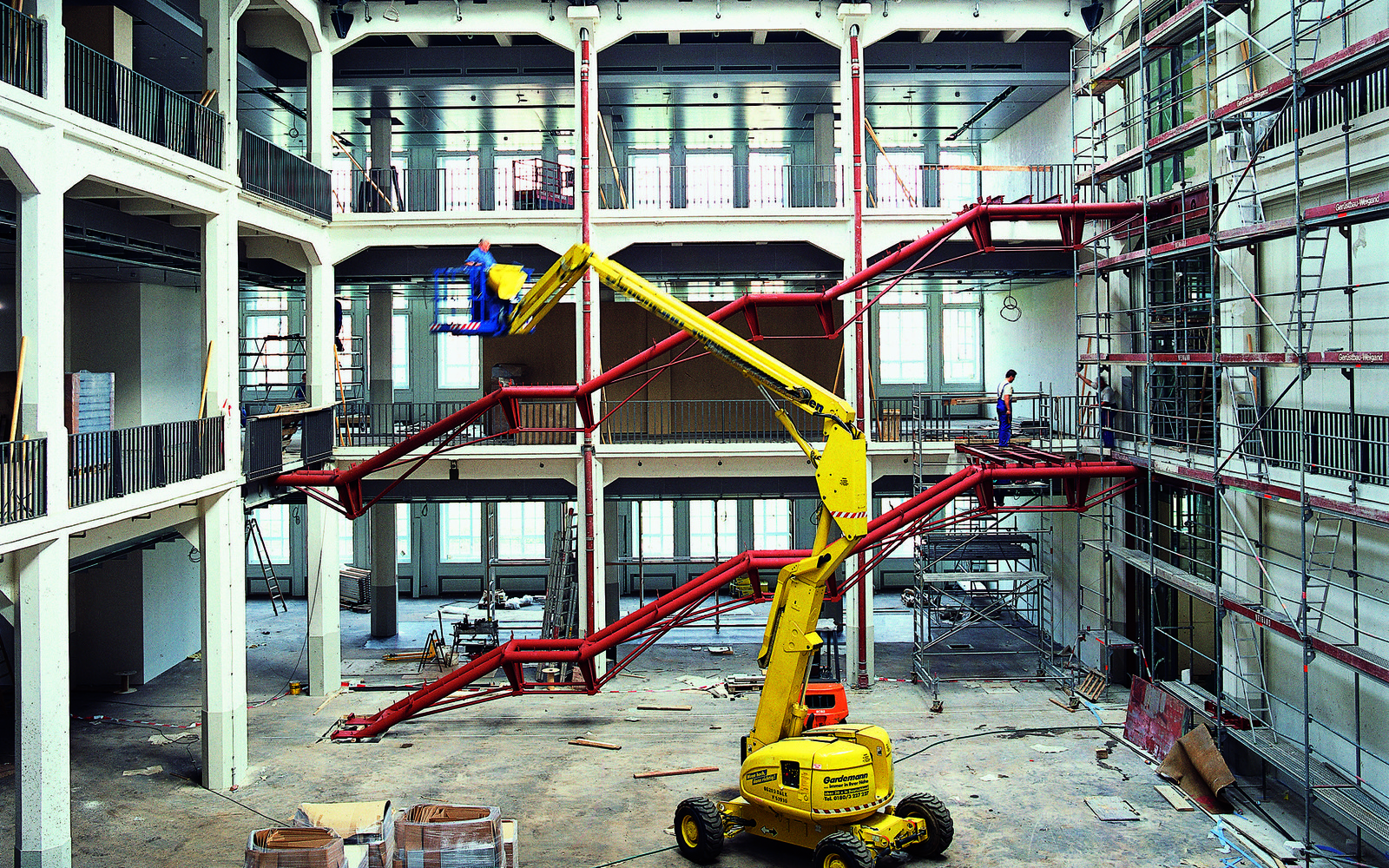 Das Foto zeigt eine historische Ansicht des Umbaus des alten MNK Hallenbaus. Ein imposanter großer gelber Bagger steht mitten in der Halle während im Hintergrund die Rohkonstruktion der zukünftigen Treppen des Ausstellungsraumes in rot hell leuchten.