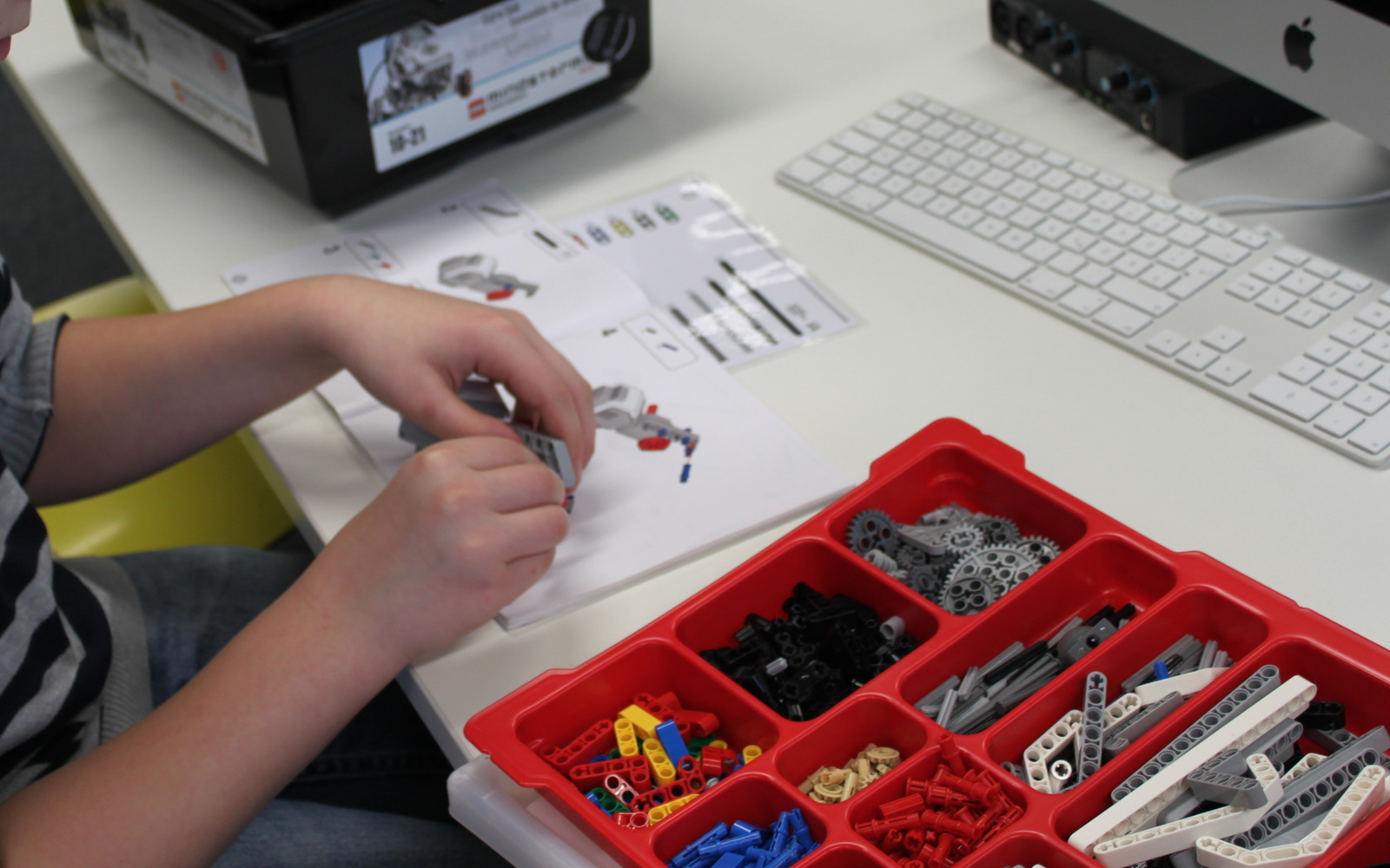 Ein Junge arbeitet an einem Lego-Roboter, den er vor sich auf dem Tisch stehen hat. Um ihn herum stehen Kisten, in denen Einzelteile liegen.