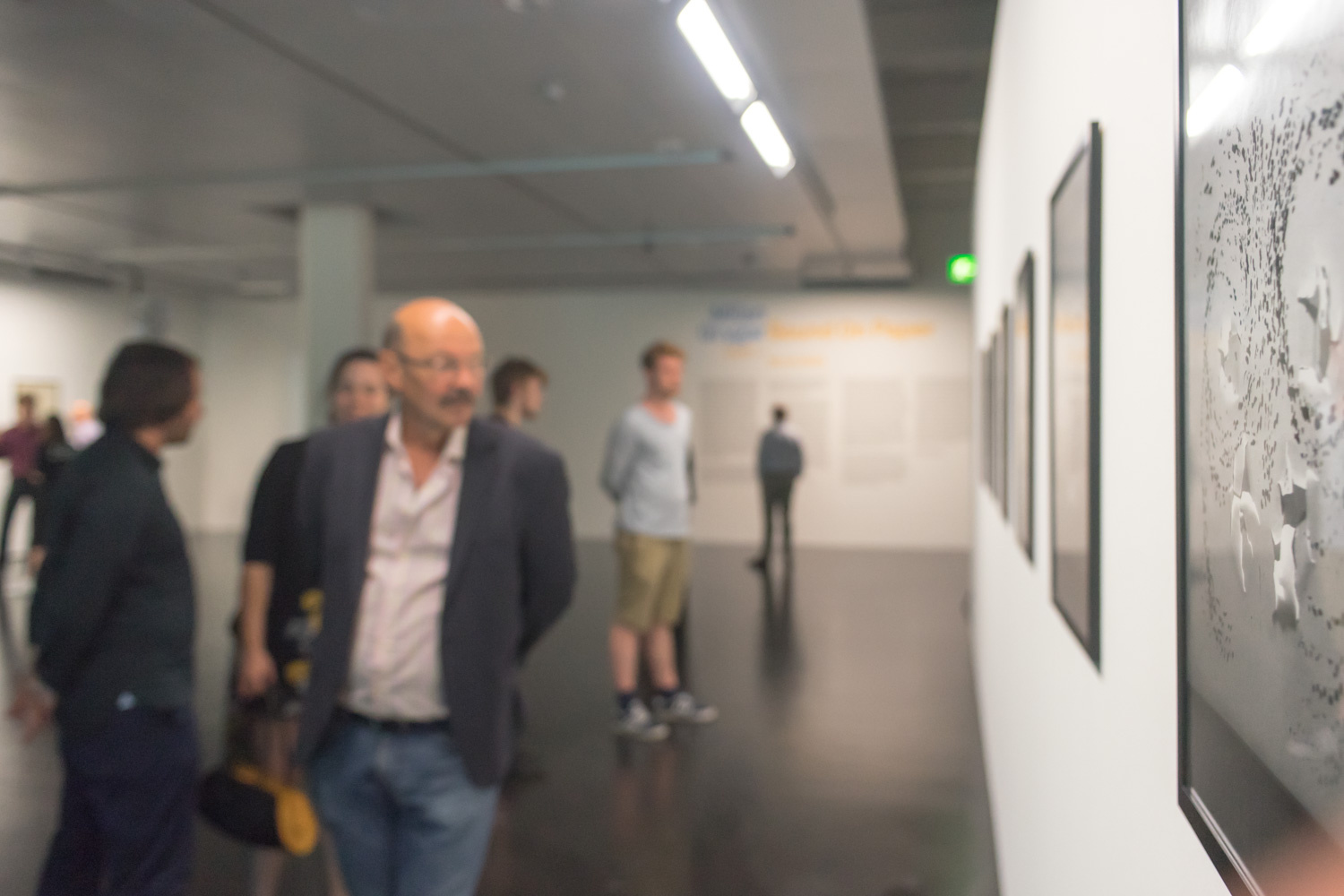 Personen im Museum bei der Ausstellungseröffnung sind nur unscharf zu erkennen. Das Bild an der Wand jedoch ist scharf und zeigt abstrakte Formen in Grautönen.