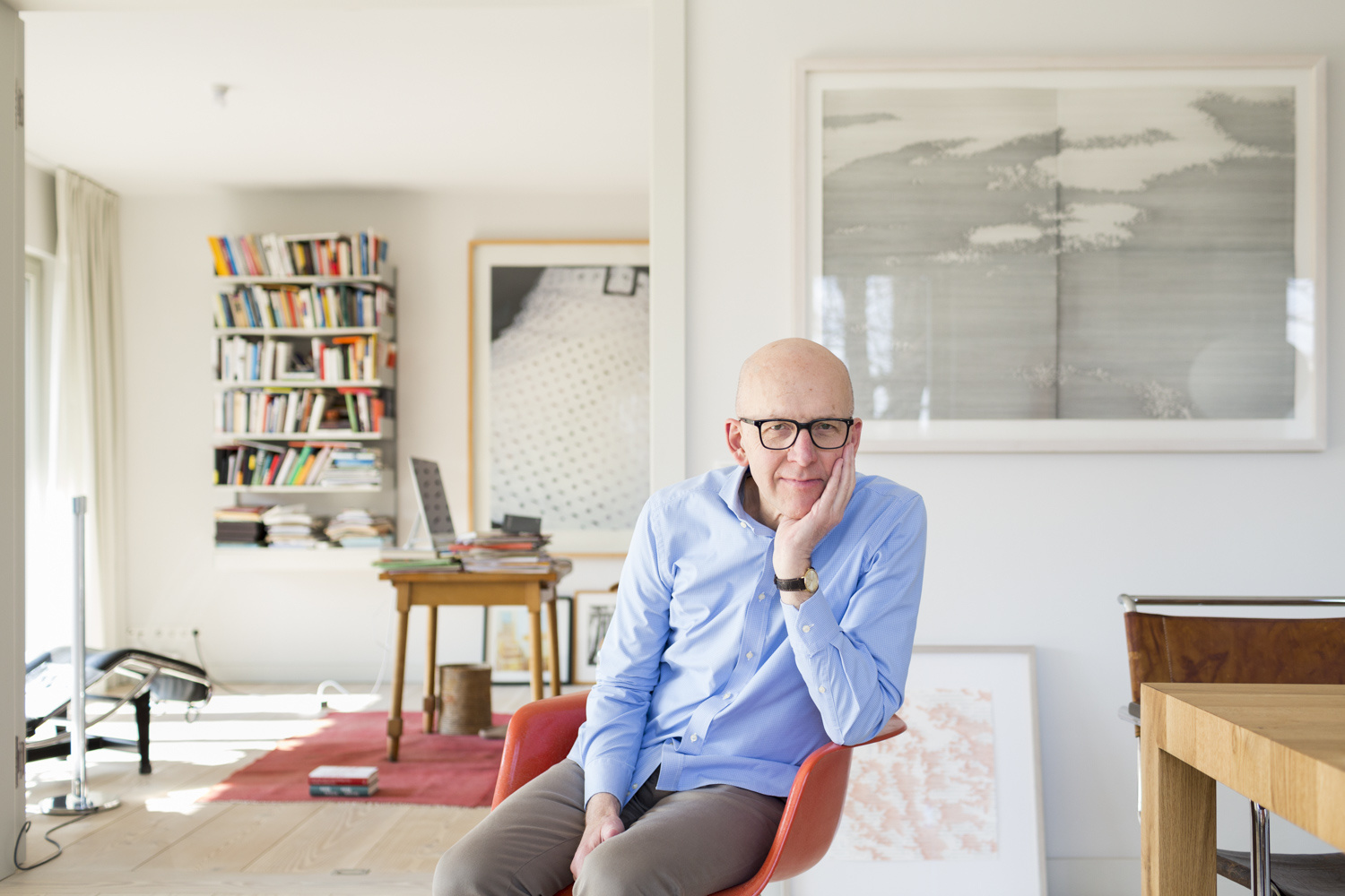 Professor Bude sits on a red chair in an apartment with bookshelf and desk, his head resting on his hand.
