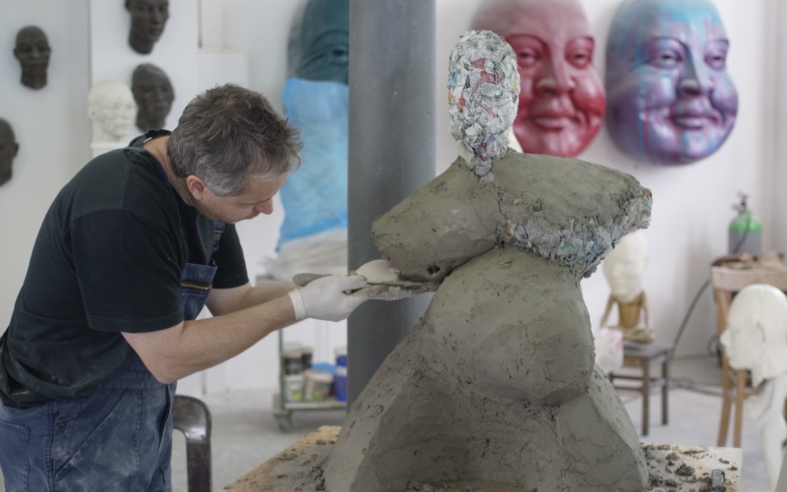 Ein Mann in Latzhosen modelliert eine voluminöse Figur aus Ton.