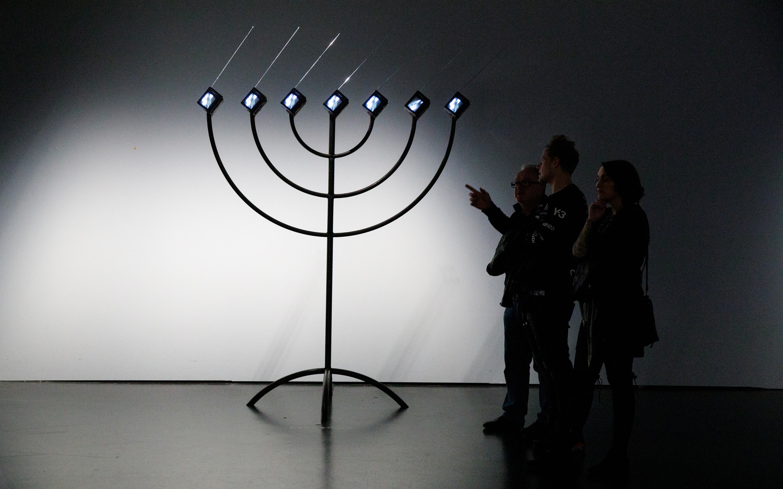 Zu sehen ist eine große, schwarze Menorah, die anstelle von Kerzen sieben kleine Bildschirme, mit nach rechts gerichteten Antennen, hat. Auf der rechten Seite stehen drei Personen, die sie betrachten. Eine der Personen zeigt auf die Installation.