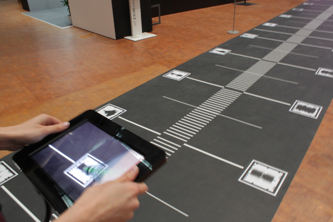 Ausschnitt eines Weges voll mit QR-Codes. Eine Hand, die mit einem iPad die QR-Codes abscannt