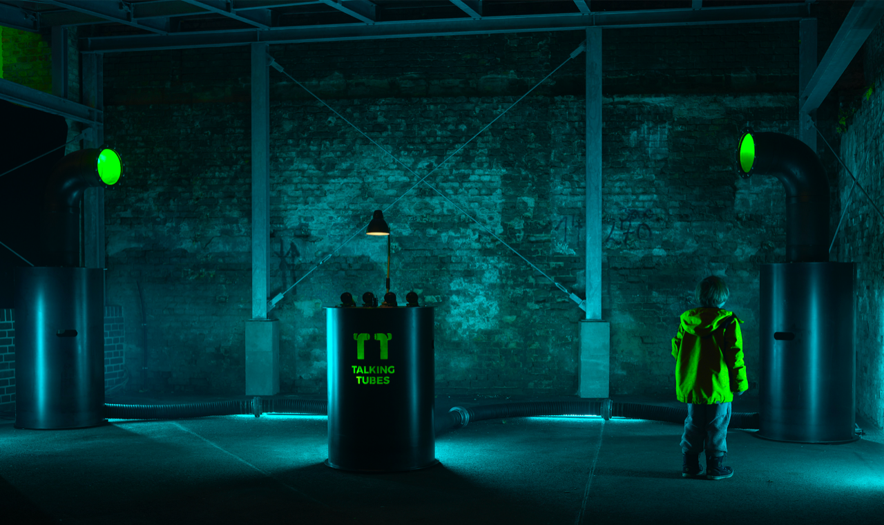 Ein blau erleuchteter Raum mit einem Kind, das die »Talking-Tubes«-Installation betrachtet: ein Steuerungspult zwischen zwei großen Röhren