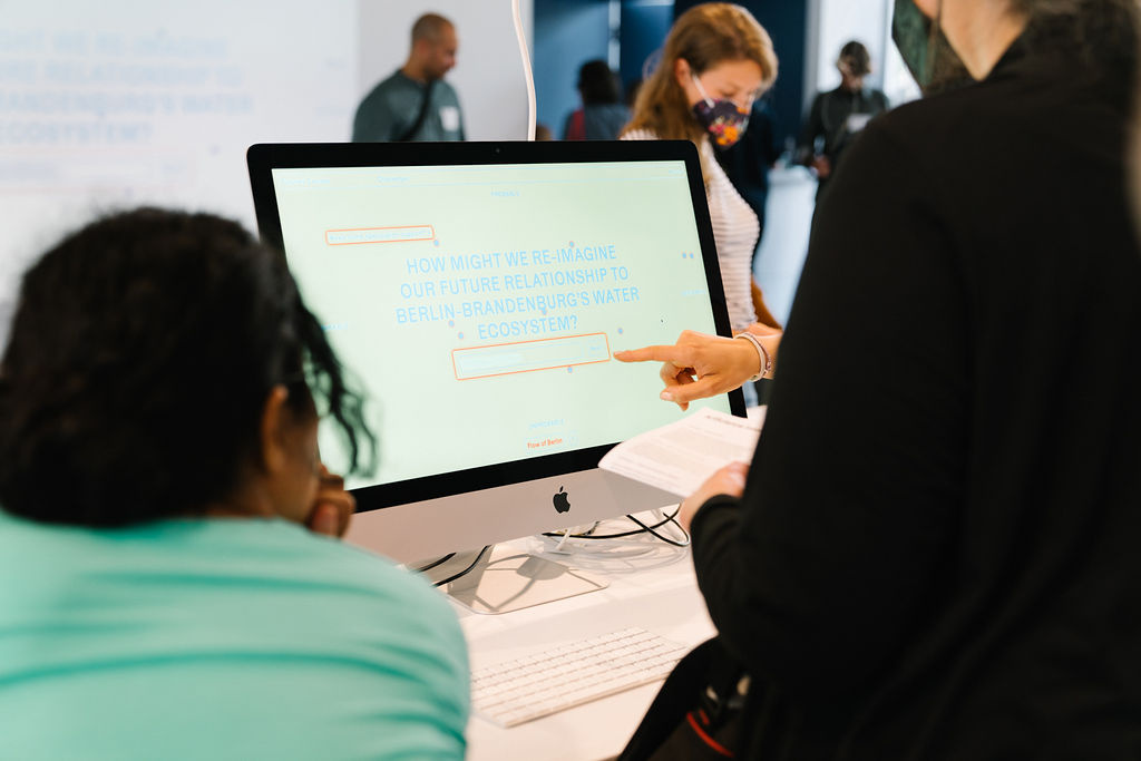 Auf dem Bild zu sehen ist ein Monitor auf dem zwei halb im Bild befindenden Menshen etwas gezeigt wird. Auf dem Monitor ist der Schriftzug "How might we're imagine our Future reationship to Berlin Brandenburg Water Ecosystem" zu lesen