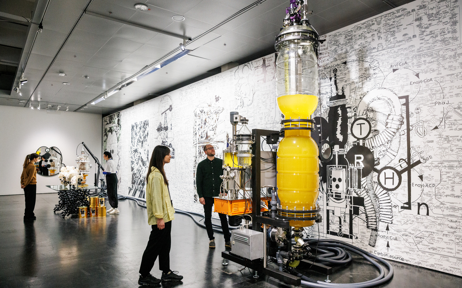 A ceiling-high, elongated glass vessel containing a yellow liquid can be seen. Several cables or hoses lead to smaller glass vessels that also contain liquid. Large hoses are lying on the floor. Two people are looking at this.