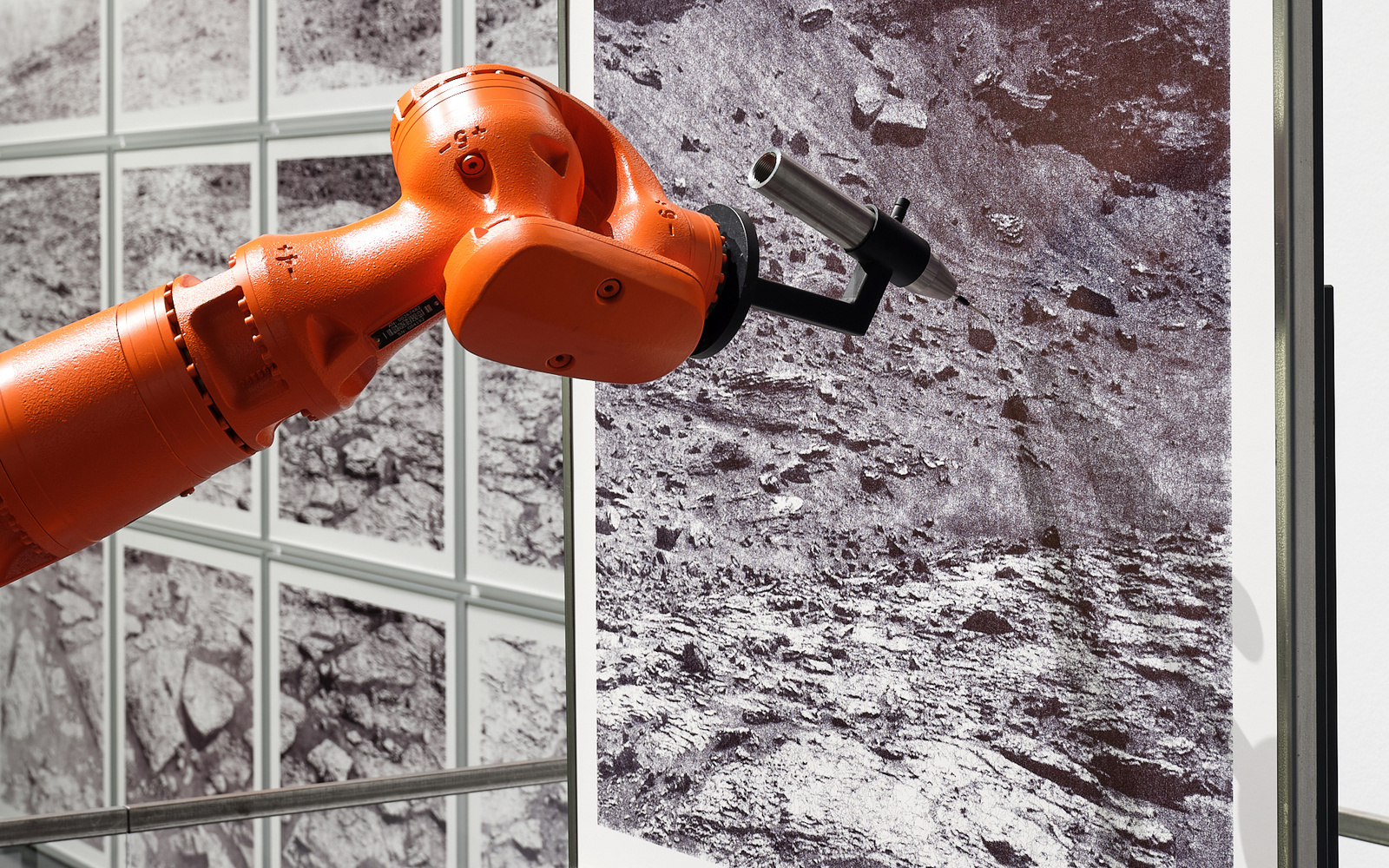 »the native picture« von robotlab. Zu sehen ist ein orangener Roboterarm, der ein schwarz-weiß Bild von einer dürren Landschaft zeichnet.