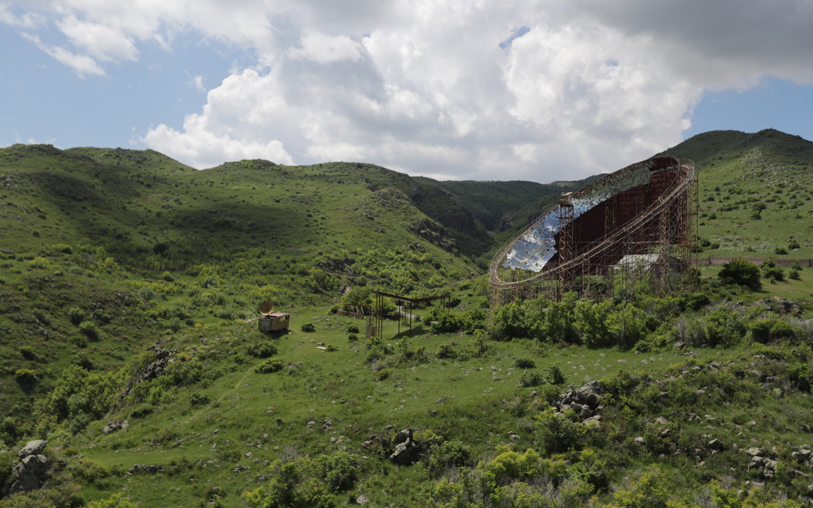 In a green, hilly landscape, a large curved metal structure with reflective surfaces stands beneath a cloudy sky.