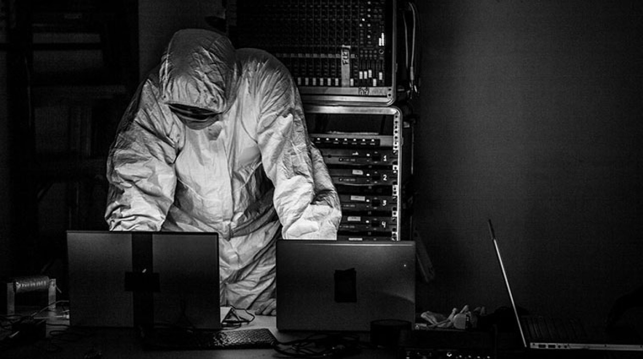 Ein Mensch in einem weissen Schutzanzug hinter einem Pult mit Laptops
