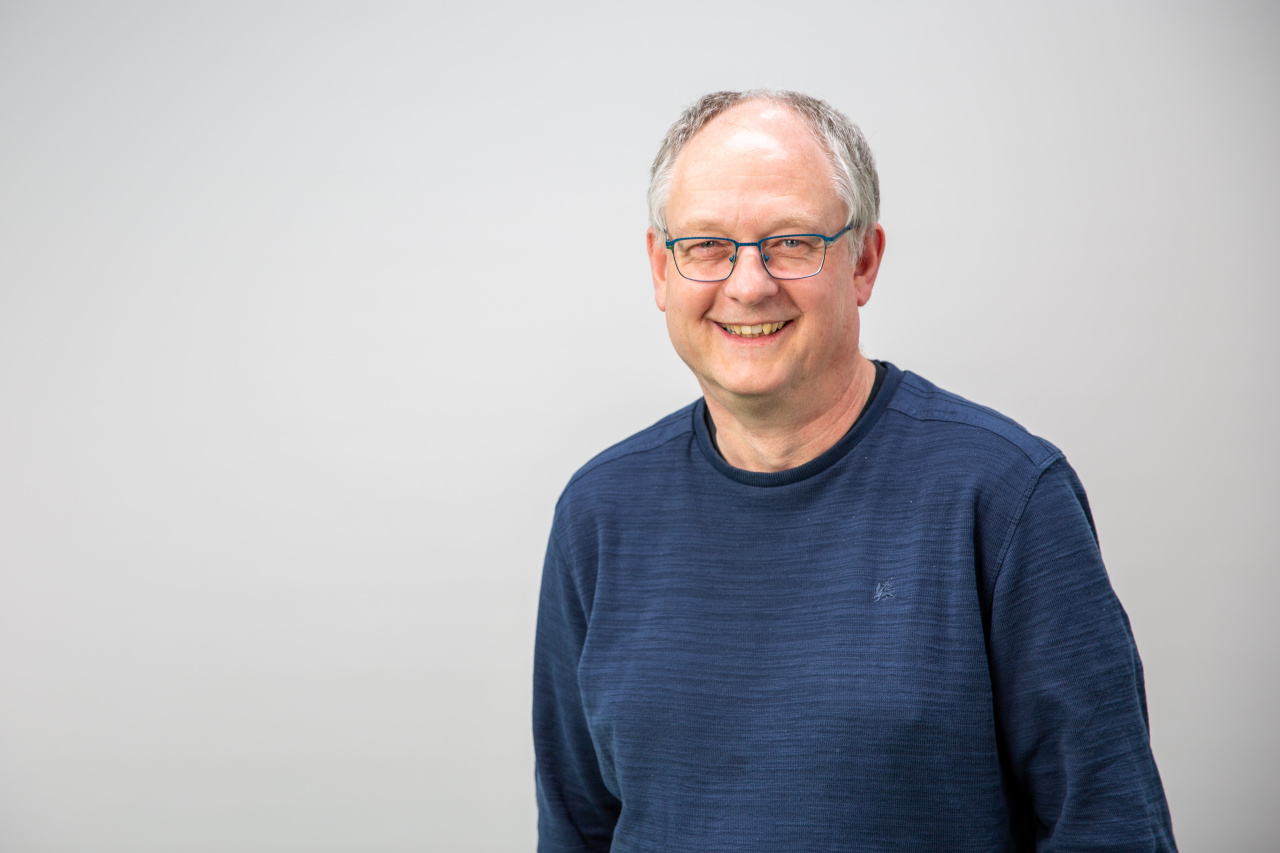 stehender Mann mit grünem Pullover, grauen Haaren und Brille
