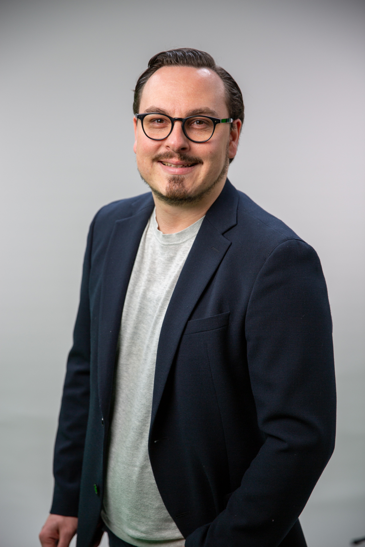 stehender Mann mit hellgrauem Pullover und einem blauen Jacket darüber