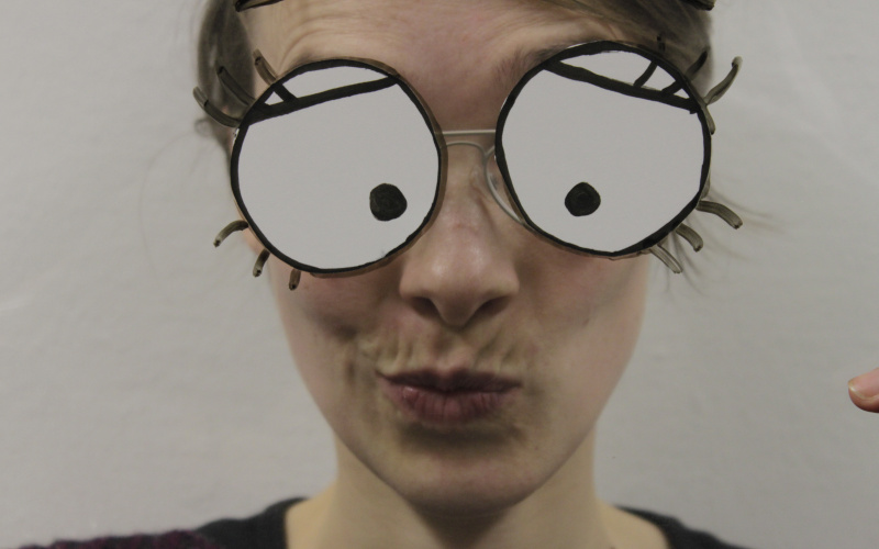 Eine junge Frau schaut verschmitzt mit gekräuselten Lippen in die Kamera. Ihre großen comic-artigen Augen sind auf eine Folie aufgezeichnet, die sie vor ihr Gesicht hält.