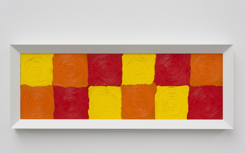 Zwölf farbig gemalte Quadrate in zwei Reihen übereinander in den Farben Gelb, Orange und Rot. Als Bild hängt es an der Wand und ist weiß gerahmt.