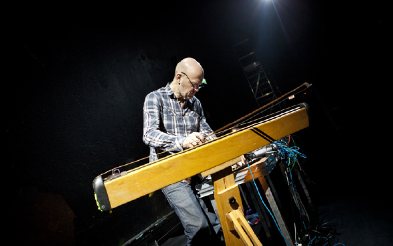 The background is black. A bespectacled man dressed in a shirt and jeans, playing an instrument and served to the mixer.
