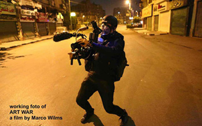 A road at night. In the foreground a man dressed in black with a helmet and protective mask. Equipment in his hands.