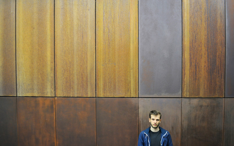 A young man wearing blue sports jacket standing in front of a wooden wall
