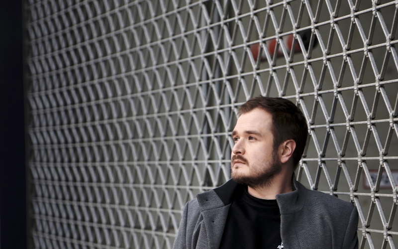 Foto von Artur Dziuk mit grauem Mantel und schwarzen Haaren. Er lehnt an einem Maschendrahtzaun.