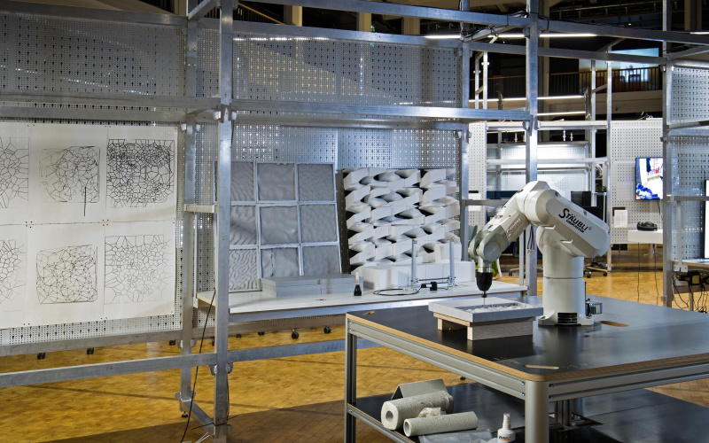 In the foreground is a robot arm on a wooden table, while in the background you can see various sculptures and drawings on a grid structure