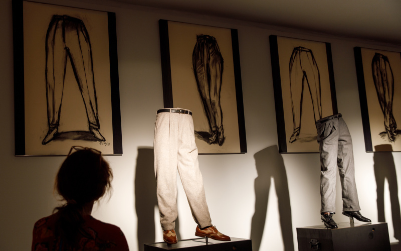 Foto einer Kunstinstallation: zu sehen sind Hosen mit Schuhen auf einer Art Laufsteg, im Hintergrund sind gerahmte Zeichnungen.