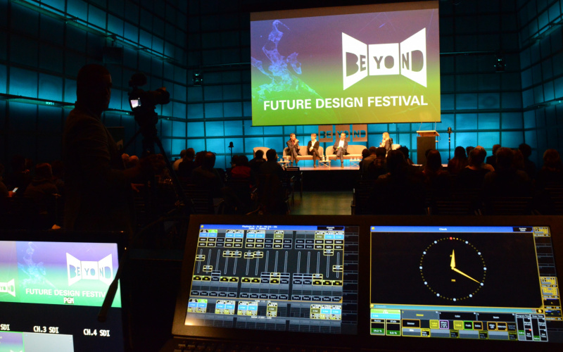 Blick in einen großen abgedunkelten Raum. An der gegenüberliegenden Wand ist eine große Leinwand mit dem Logo des Beyond Future Design Festivals zu sehen.