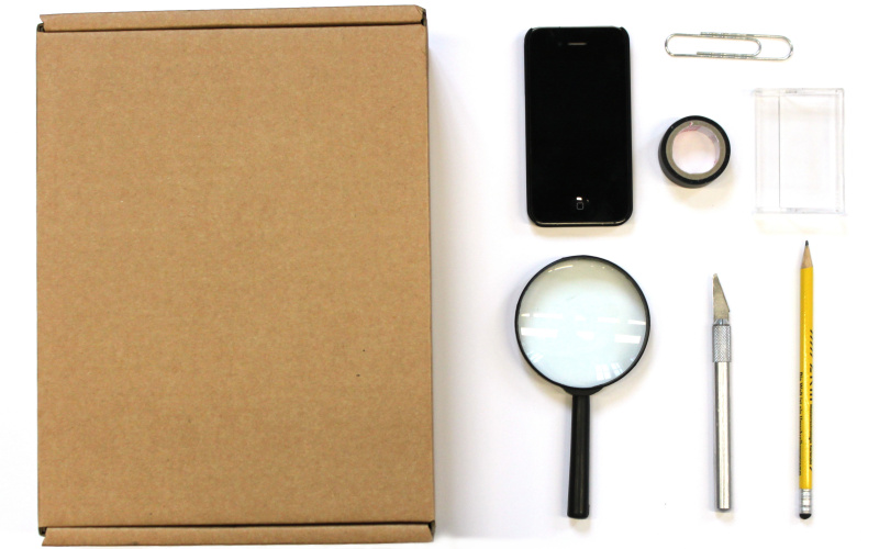 Zu sehen ist eine Lupe, ein Smartphone, eine Pappkiste, ein Cutter, ein Stift, eine Büroklammer, eine Kassettenhülle und Klebeband.