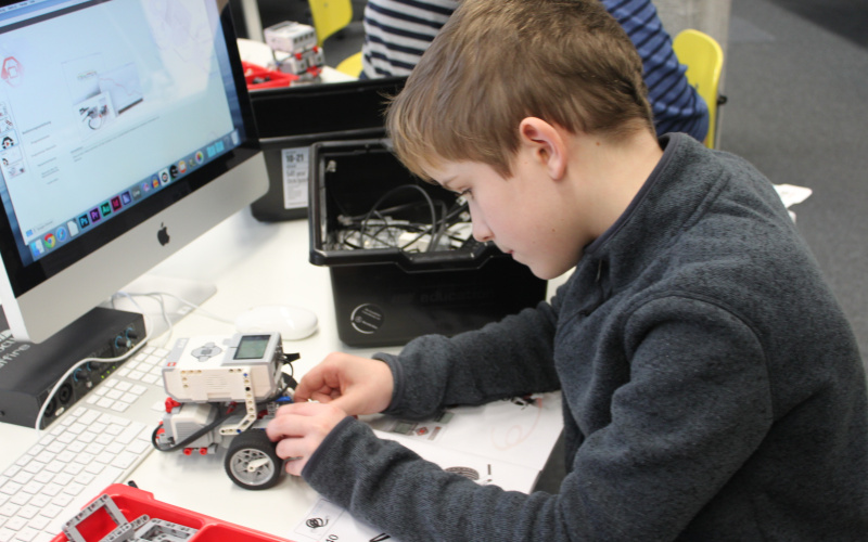 Ein Junge arbeitet an einem Lego-Roboter, den er vor sich auf dem Tisch stehen hat. Um ihn herum stehen Kisten, in denen Einzelteile liegen.