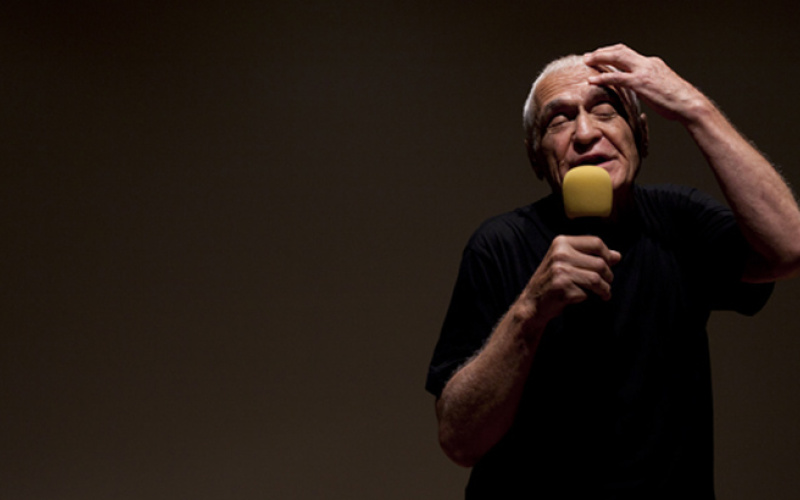 A man speaks into a microphone and holds up his left hand to his head