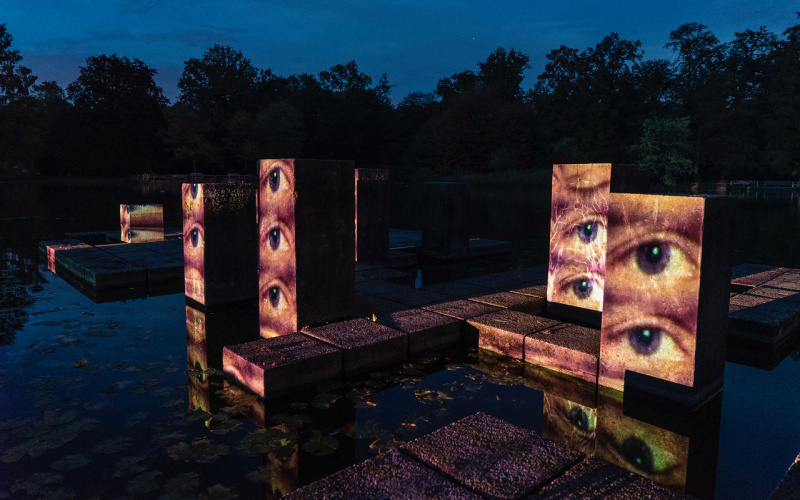 Wasserstelen in einem Park. Es ist später Abend. Auf die Stelen sind Augen gemappt, die offen in die Gegend blicken.