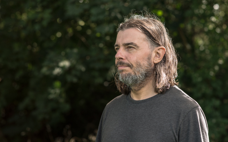 You can see the portrait of a bearded middle-aged man with long hair. He stands in front of a tree and looks past the camera. 