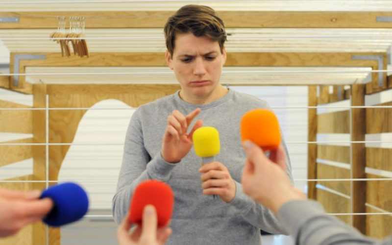 Ein junger Mann tippt kritisch mit seinem Finger auf ein gelbes Mikrofon. Er ist von verschiedenen Mikrofonen umgeben.
