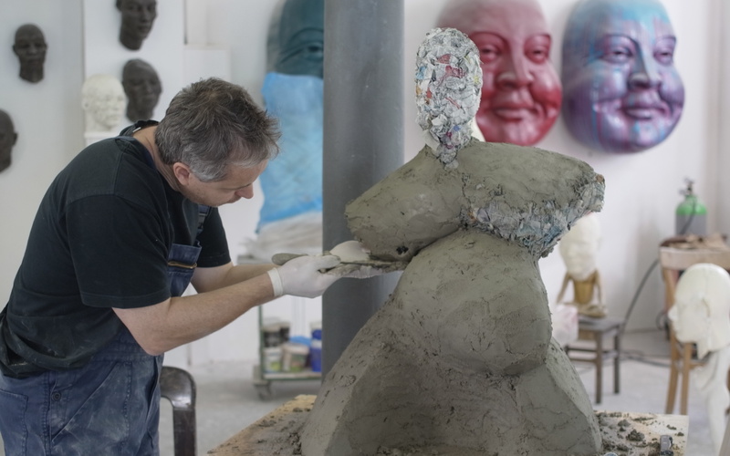 Ein Mann in Latzhosen modelliert eine voluminöse Figur aus Ton.