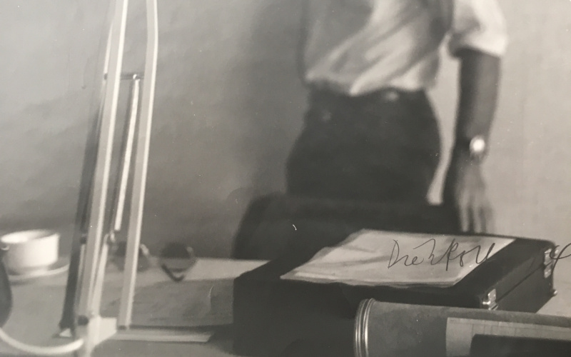 Eine Schwarz Weiß Fotographie zeigt im Vordergrund scharf eine Schreibtischlampe. Im Hintergrund sind man unscharf einen Mann mit Bart und einem weißen Hemd. Es ist Hansjörg Mayer.