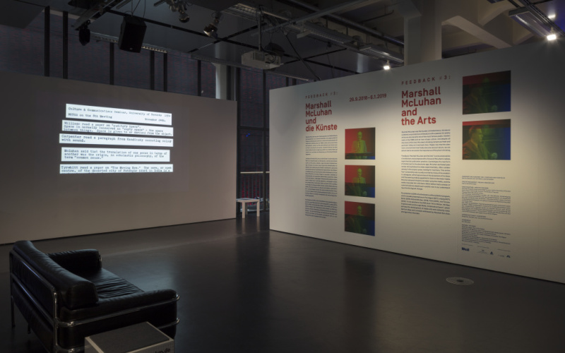 Im Vordergrund auf der linken Seite steht ein schwarzes Sofa. Hinten links steht eine Stellwand mit Text-Projektion. Hinten rechts eine mit Text und Bildern bedruckte.