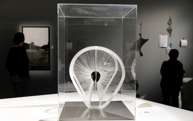 Ein schwungvoll gearbeitetes Kunststoffband ist von zahlreichen Fäden umspannt. Die Fäden sind radial angeordnet wie die Speichen eines Fahrrads. 